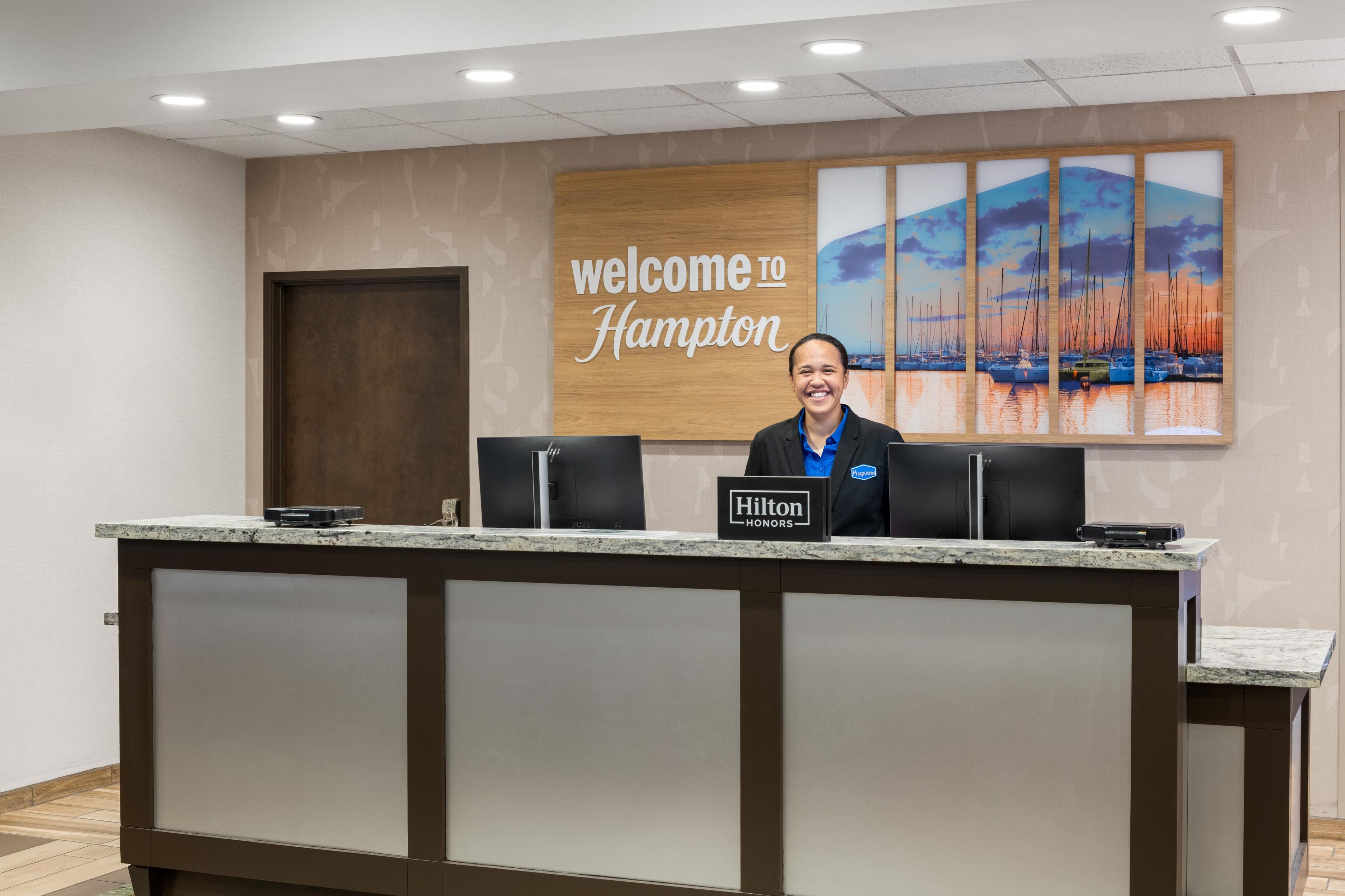 Un comptoir d’accueil d’hôtel avec une pancarte « Bienvenue à Hampton », une plaque nominative pour « Hilton Honors » et un décor représentant des voiliers.