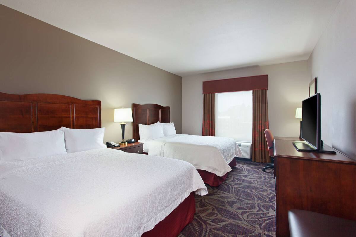 A hotel room with two beds, a desk, a television, and a window with curtains.