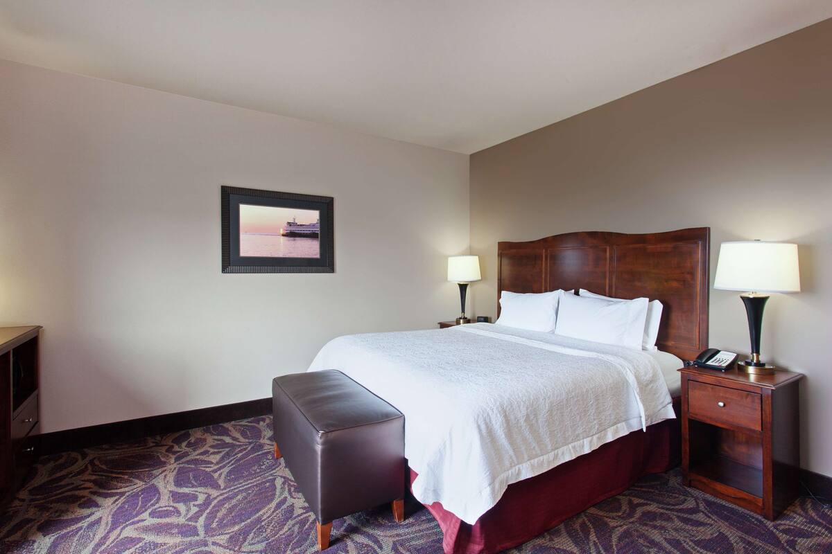 A hotel room with a large bed, two bedside tables with lamps, a small wooden bench, and a framed picture on the wall.