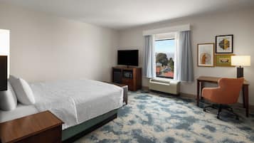 Ein Hotelzimmer mit Bett, Fernseher, Schreibtisch mit Stuhl, Fenster mit Vorhängen und gerahmten Bildern an der Wand.