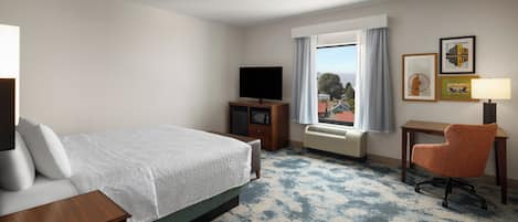 A hotel room with a bed, a television, a desk with a chair, a window with curtains, and framed pictures on the wall.
