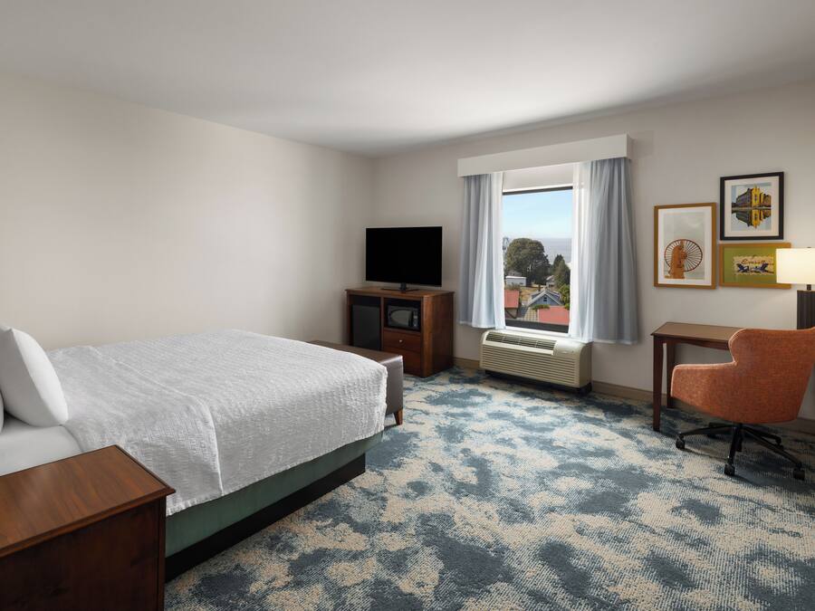 A hotel room with a bed, a television, a desk with a chair, a window with curtains, and framed pictures on the wall.