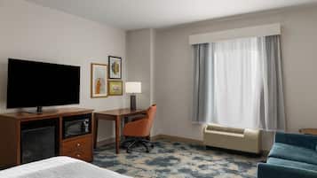 Habitación de hotel con cama, escritorio, televisión, silla, lámpara, ventana con cortinas y calefacción.