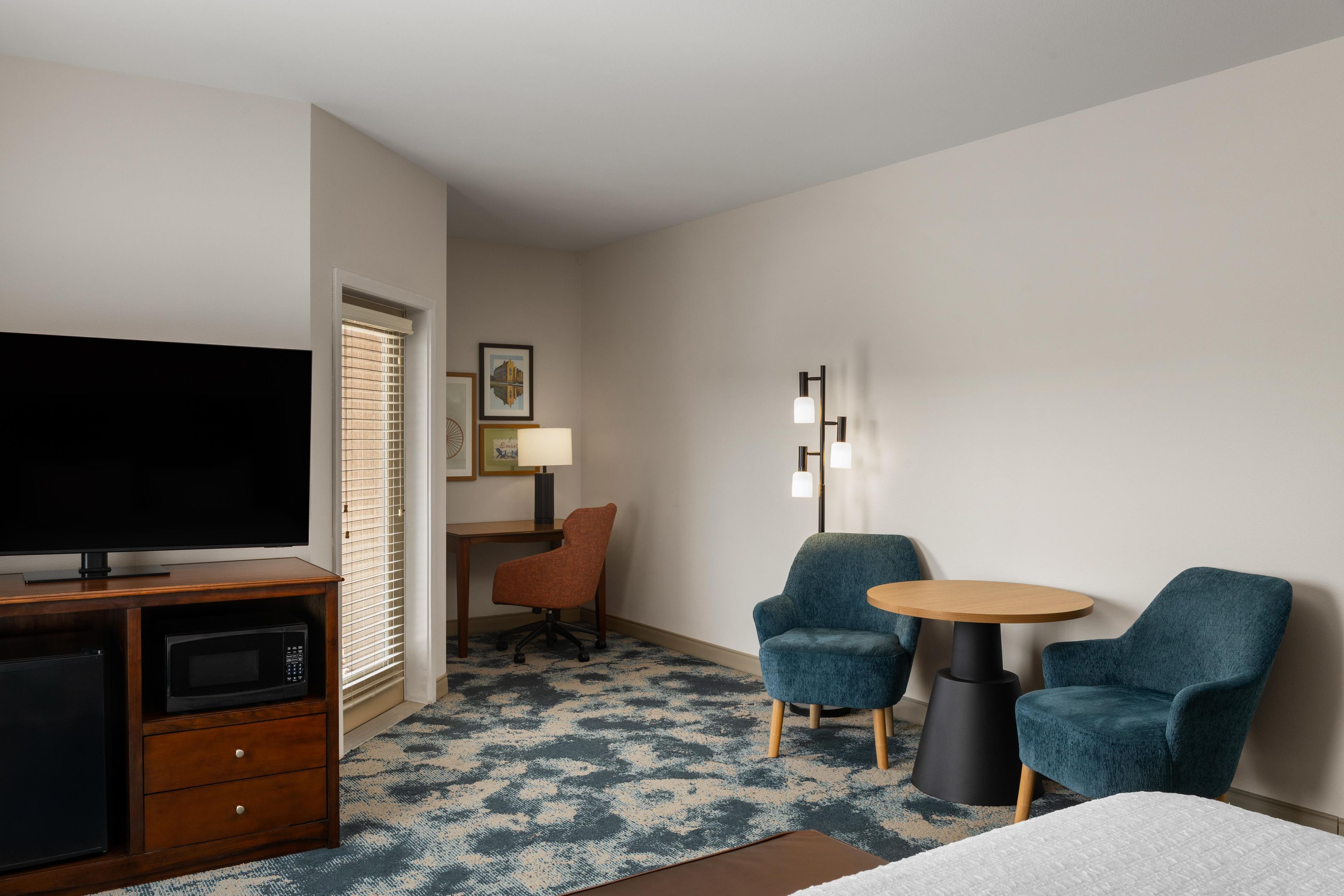 A hotel room with a flat-screen TV, a desk with a lamp, two armchairs, a small table, and a patterned carpet.