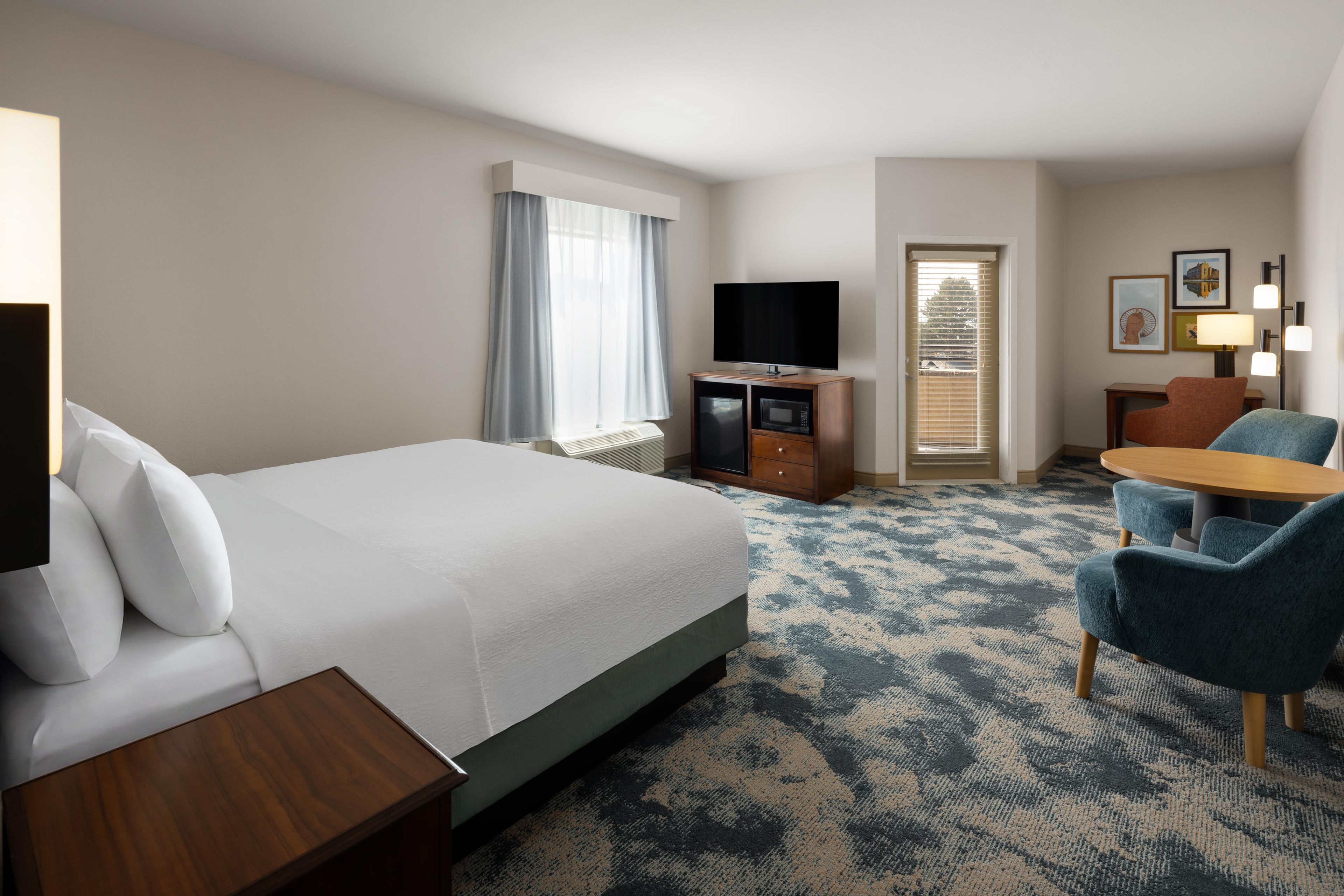 A hotel room with a large bed, a desk, a television, a seating area with chairs, and a window with curtains.