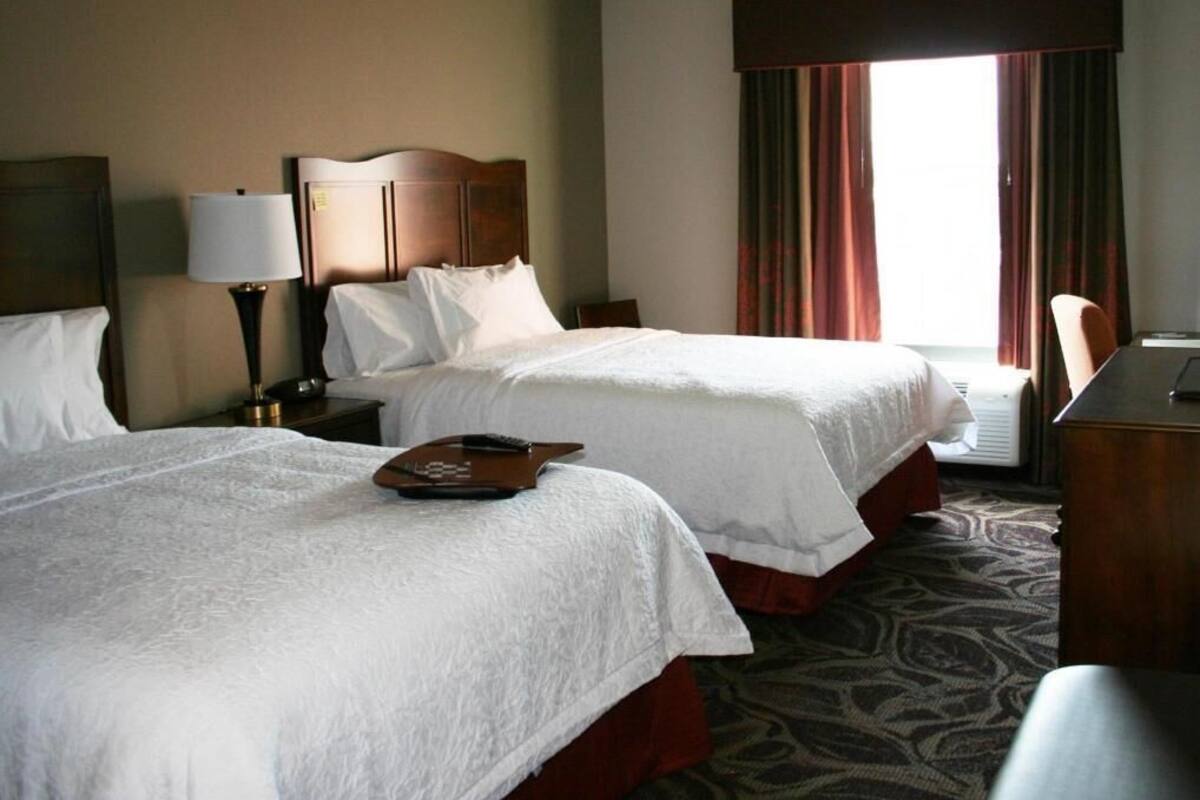 A hotel room with two beds, a desk, a lamp, and a window with curtains.