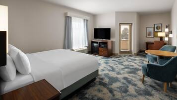 A hotel room with a large bed, a desk, a television, a seating area with chairs, and a window with curtains.