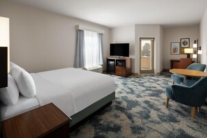 Habitación de hotel con una cama grande, un escritorio, un televisor, una zona de estar con sillas y un ventanal con cortinas.
