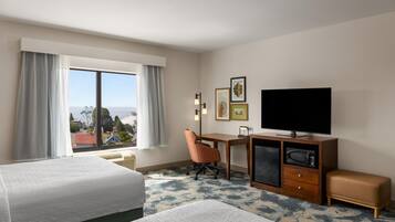 Ein Hotelzimmer mit zwei Betten, einem Schreibtisch mit Stuhl, einem Fernseher und Blick auf die Stadt.