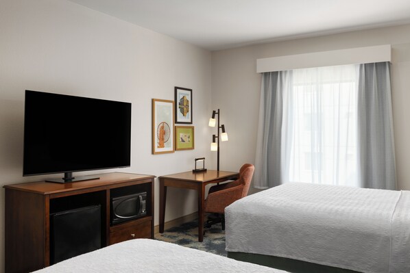 A hotel room with a flat-screen TV, a desk with a chair, two beds, a window with curtains, and artwork on the wall.
