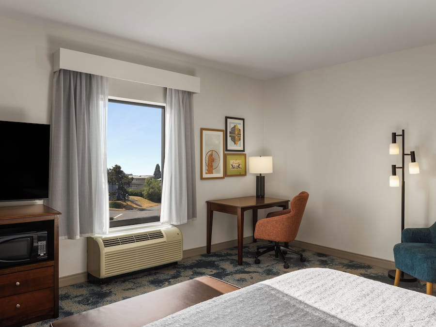 Ein Hotelzimmer mit Bett, Schreibtisch, Stuhl, Fernseher und einem Fenster mit Vorhängen.