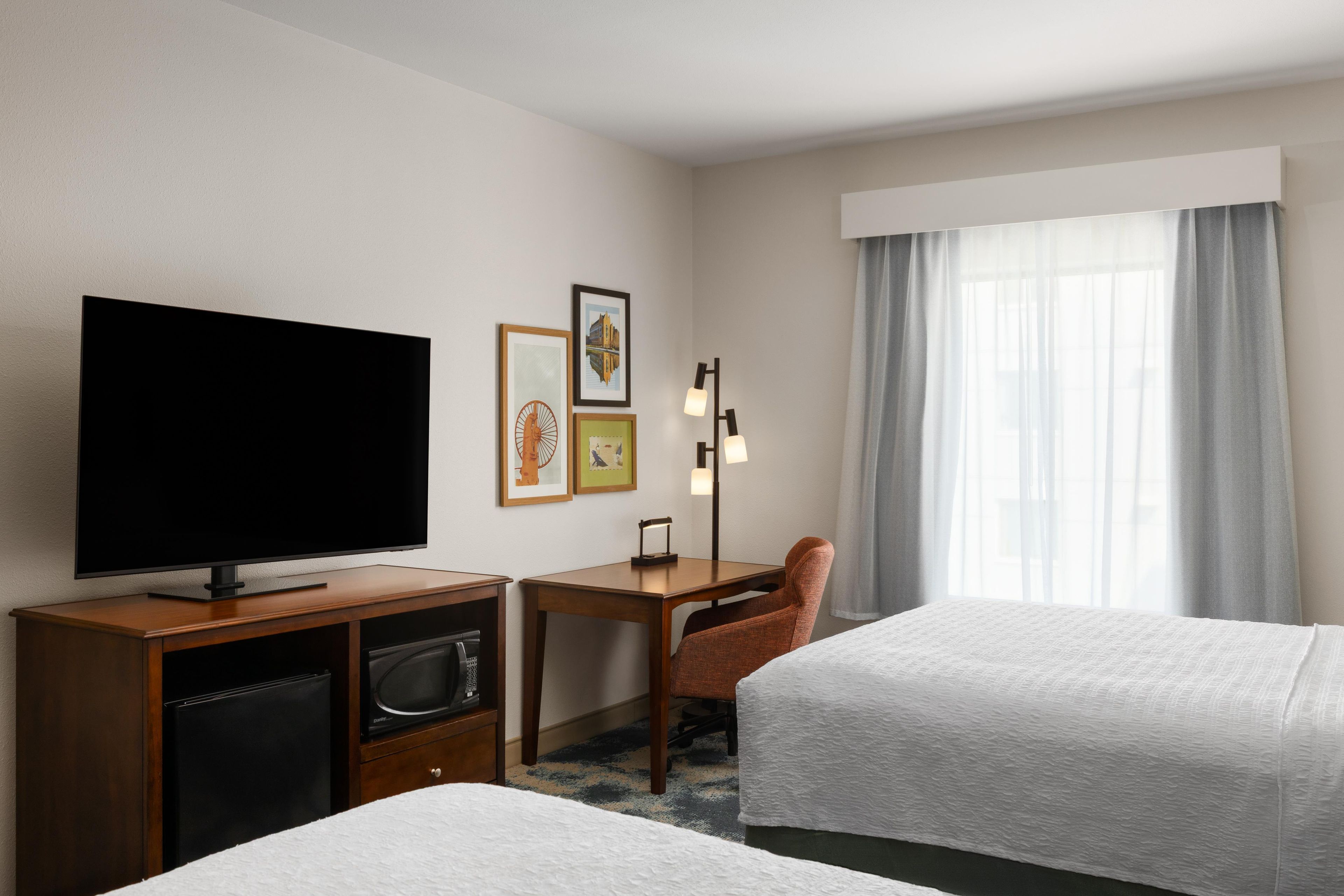 Une chambre d’hôtel équipée d’une télévision à écran plat, d’un bureau avec une lampe, de deux lits et d’une grande fenêtre avec des rideaux.