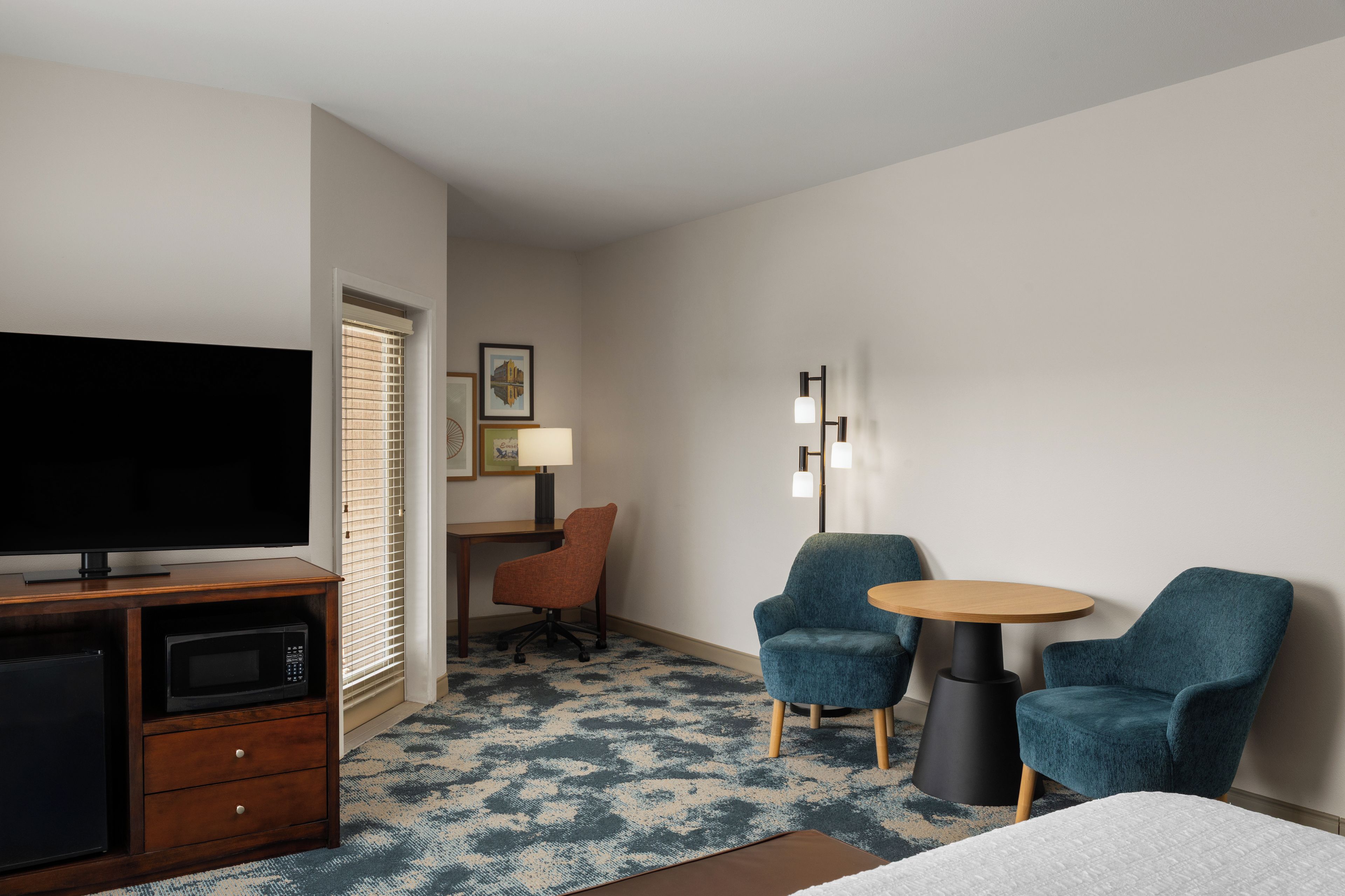 A hotel room with a flat-screen TV, a desk, two chairs, a lamp, and a floor lamp.