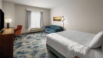 A hotel room with a bed, a desk with a lamp, a chair, a sofa, and a painting on the wall.