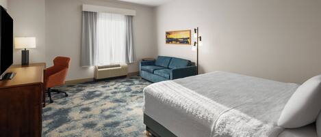 Habitación de hotel con cama, escritorio con lámpara, sillón, sofá y un cuadro en la pared.