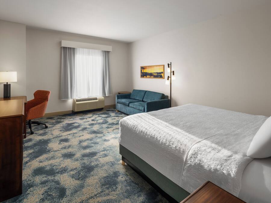 A hotel room with a bed, a desk with a lamp, a chair, a sofa, and a painting on the wall.
