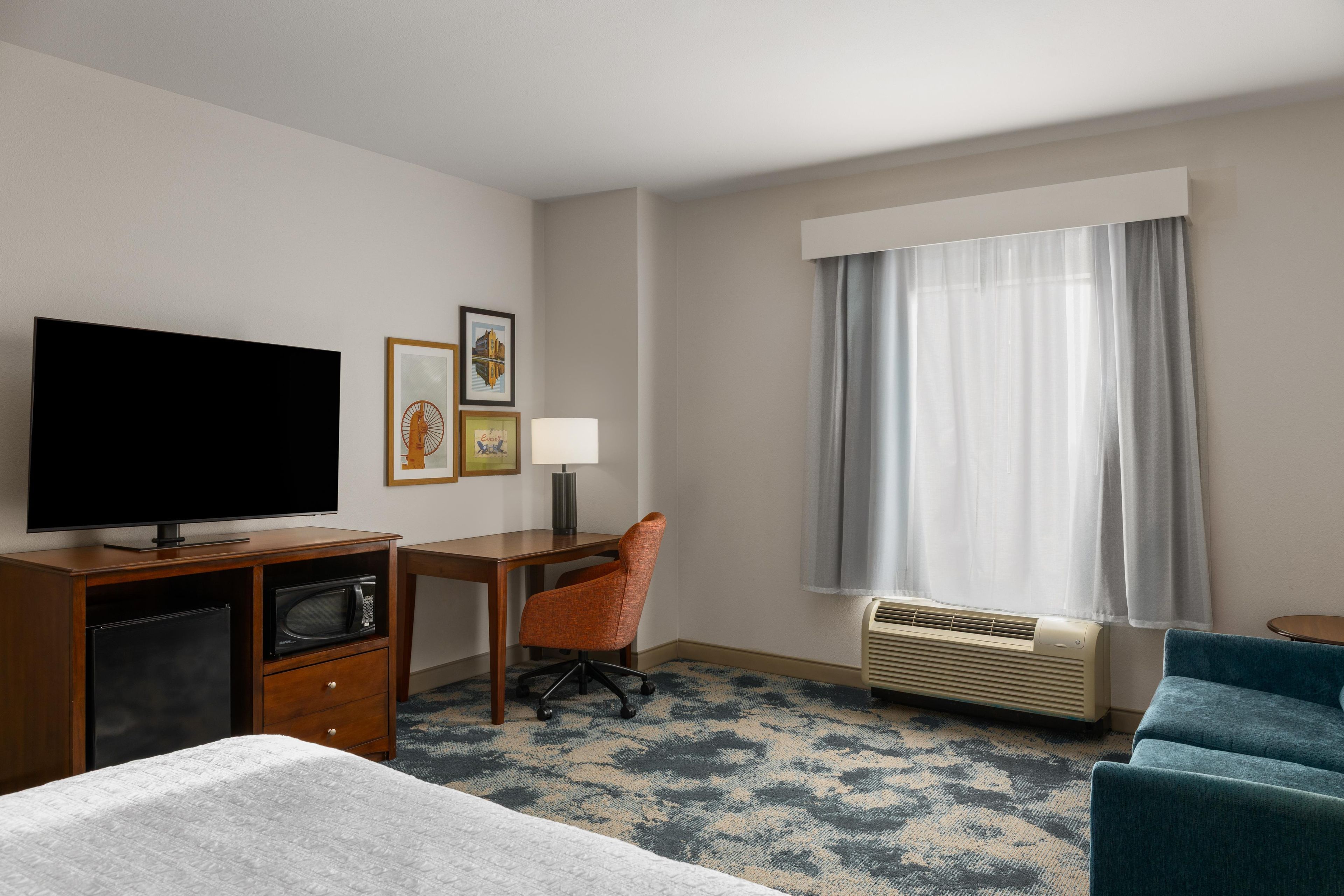 A hotel room with a bed, a desk, a television, a chair, a lamp, a window with curtains, and a heater.