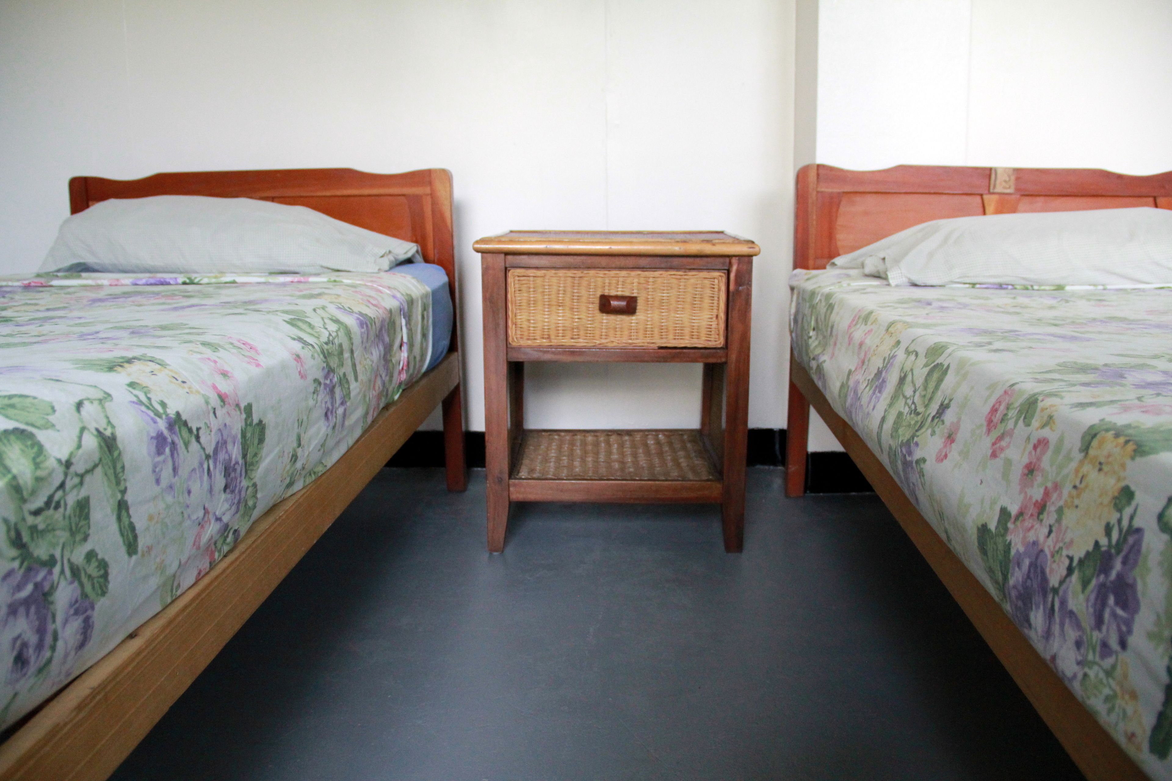 A room with two single beds, each with a wooden headboard and floral bedspread. There is a small wooden nightstand with a wicker basket between the beds.