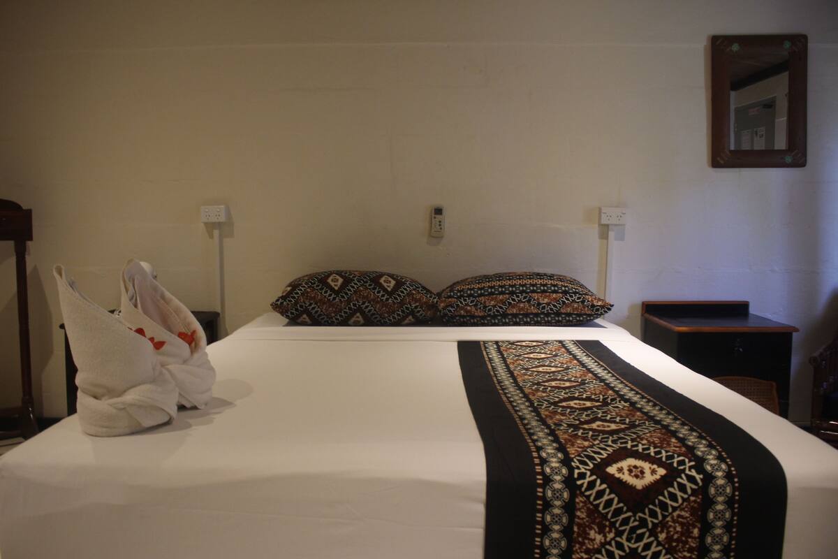 Ett hotellrum med en väl bäddad säng, två kuddar, ett sängbord, en liten avlastningsyta och en spegel som hänger på väggen.