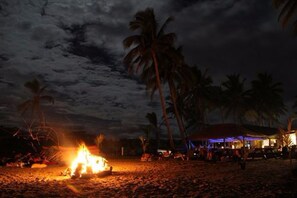 A beach scene with a campfire, palm trees, and a building with lights.