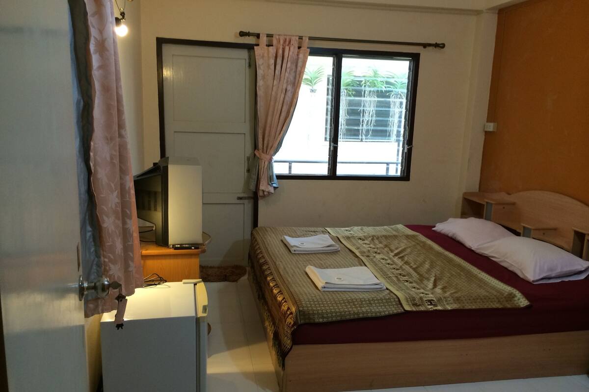 A bedroom with a bed, a small desk with a lamp, a television, and a window with curtains.