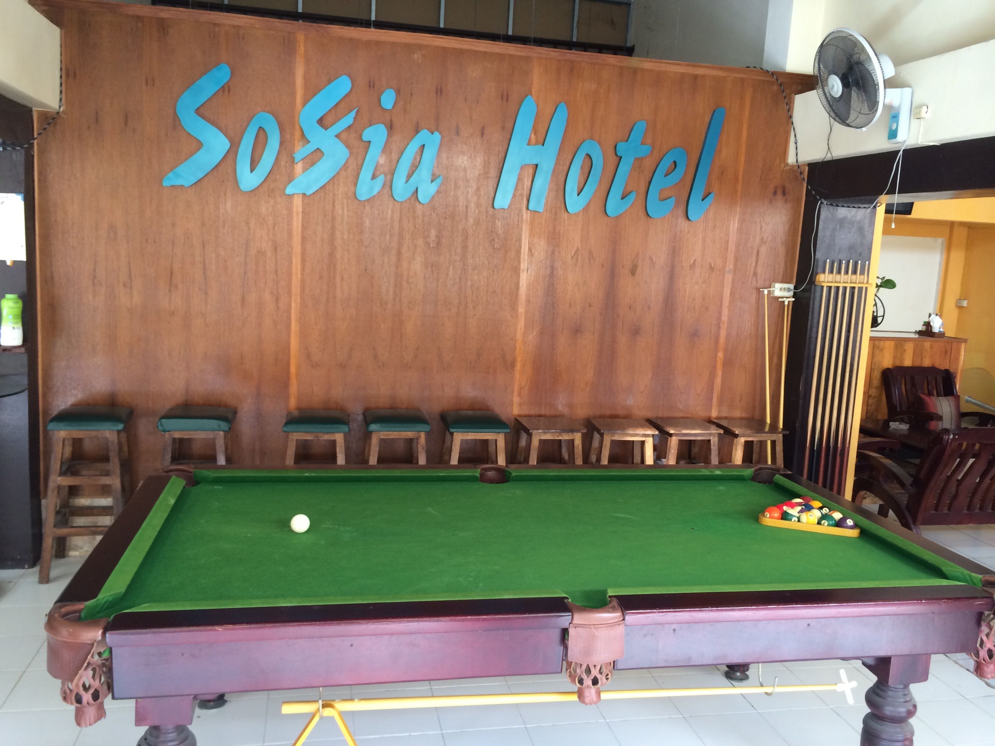 A pool table with a green felt surface, a cue ball, and a set of pool balls. There are six wooden stools aligned against a wooden wall with the text "Sosia Hotel" in blue letters.