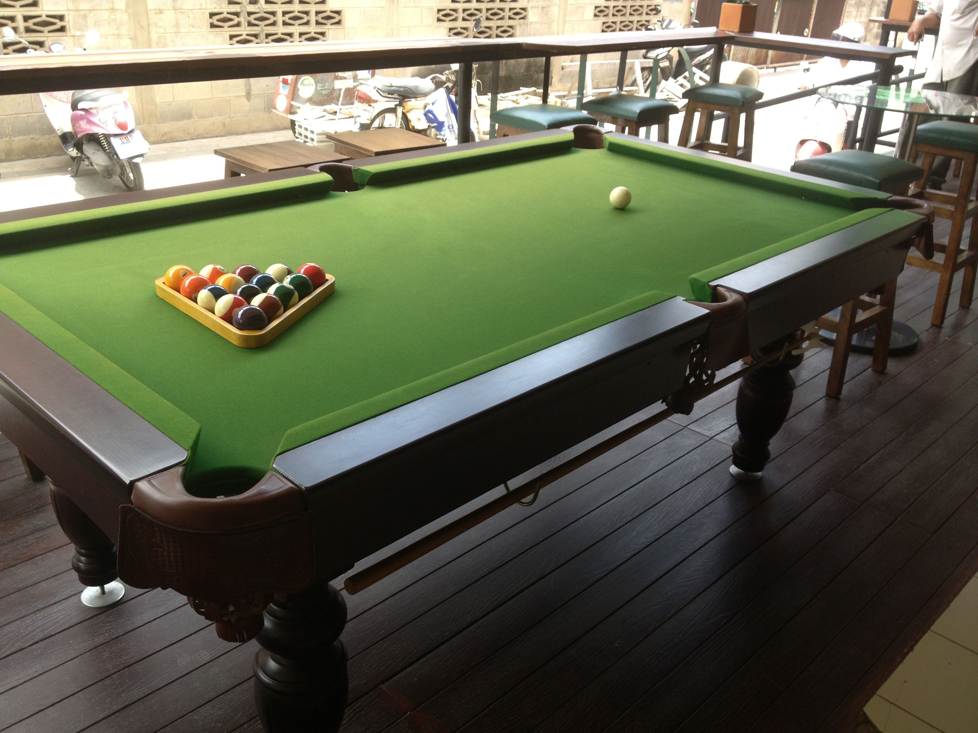 A pool table with a green felt surface, surrounded by wooden chairs and a railing.