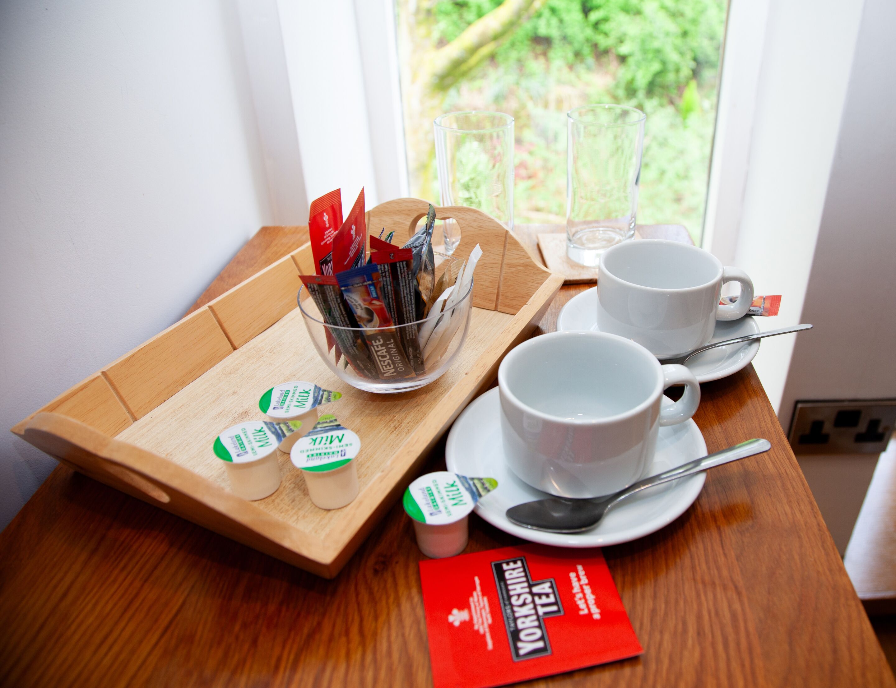 Un vassoio per la colazione con Yorkshire Tea, Nescafe Original e confezioni di latte su un tavolo di legno vicino a una finestra.