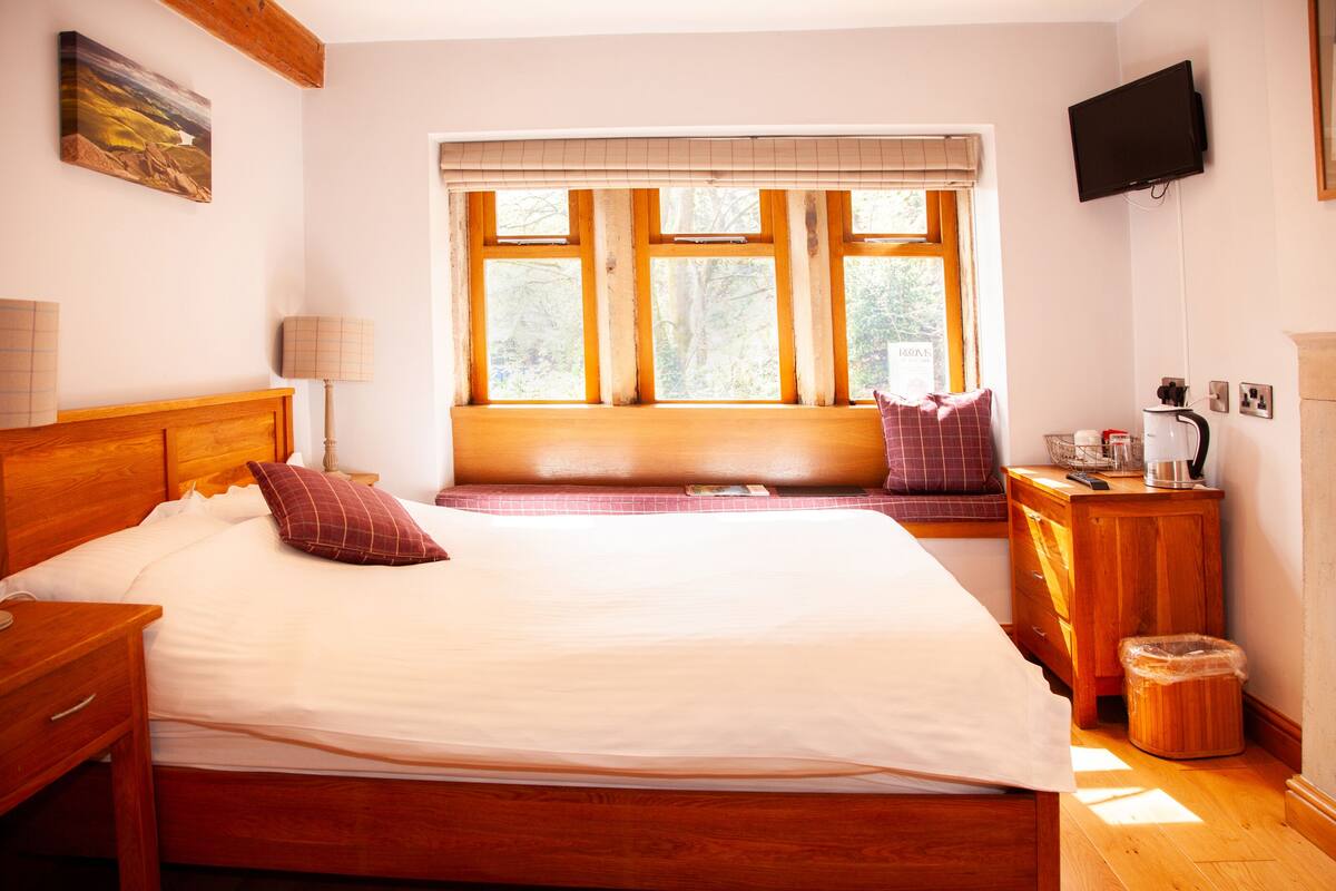 A bedroom with a large bed, a wooden headboard, a bedside table with a lamp, a window with blinds, a television mounted on the wall, and a small wooden cabinet.