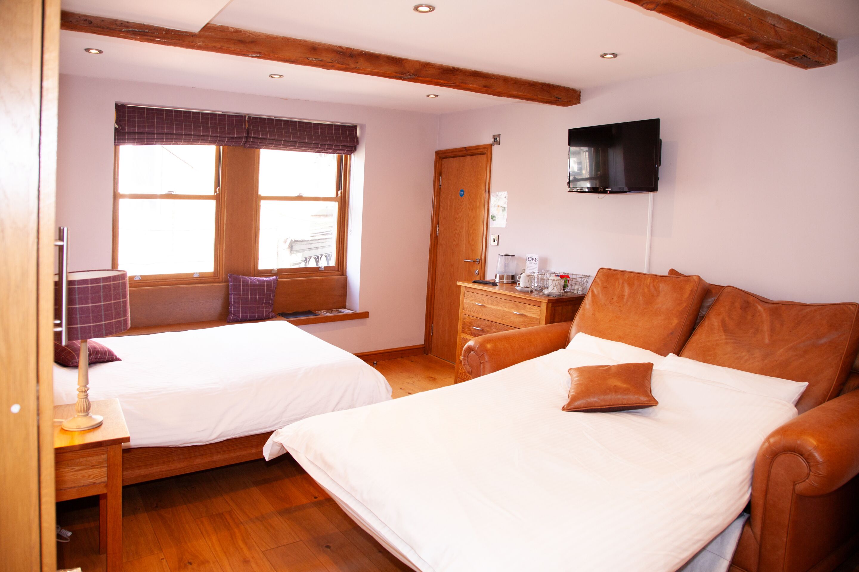 A bedroom with two beds, wooden floors, a wooden dresser, a television on the wall, and a window with blinds.