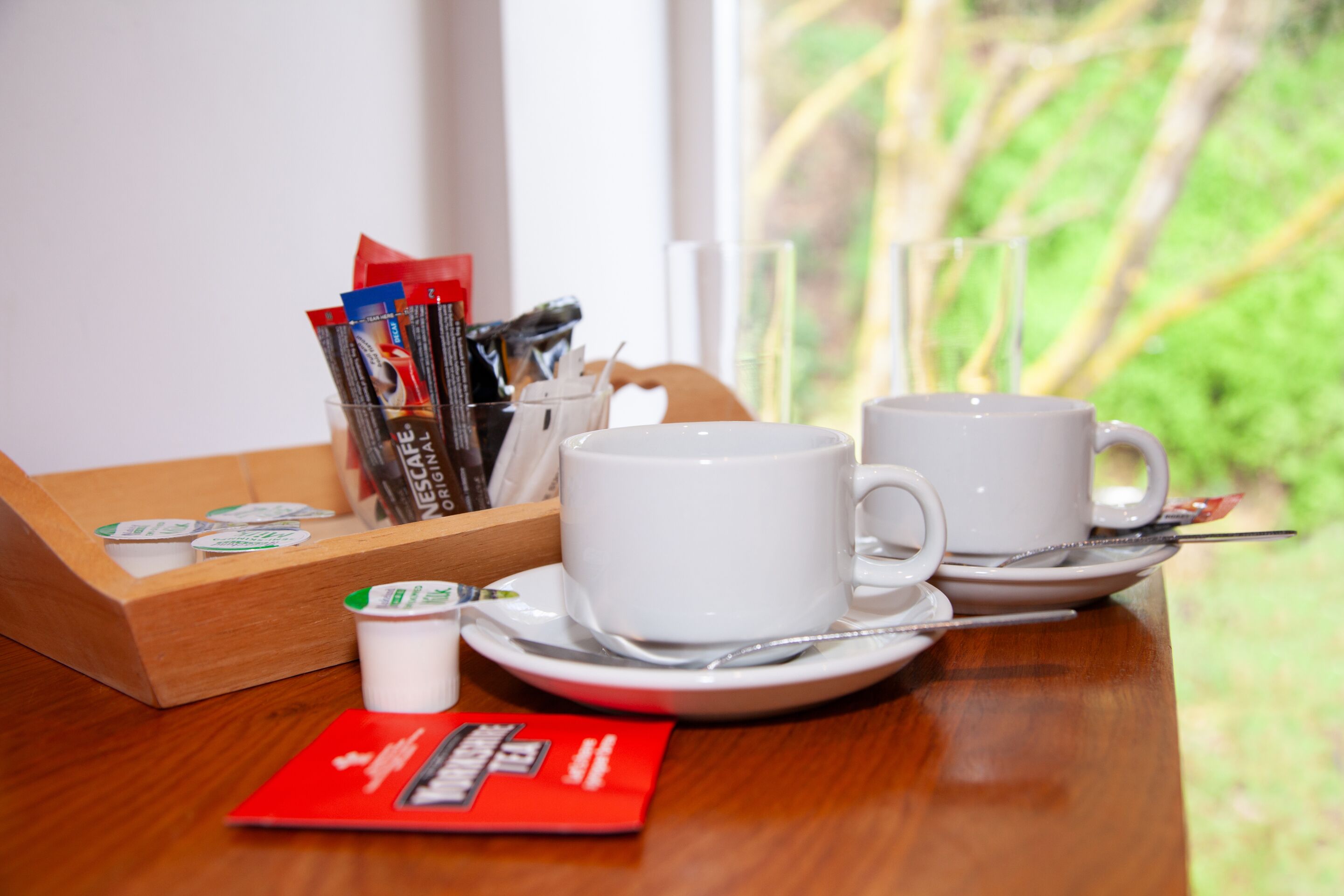 Un desayuno con dos tazas blancas sobre platillos, una bandeja con sobres de café, un vasito de crema y un folleto rojo sobre una mesa de madera.