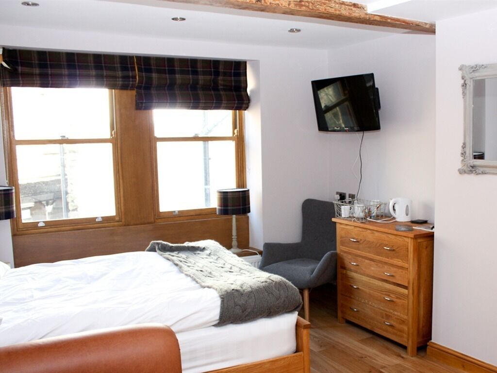 Un dormitorio con cama, una silla, cómoda, televisor y ventana con cortinas.
