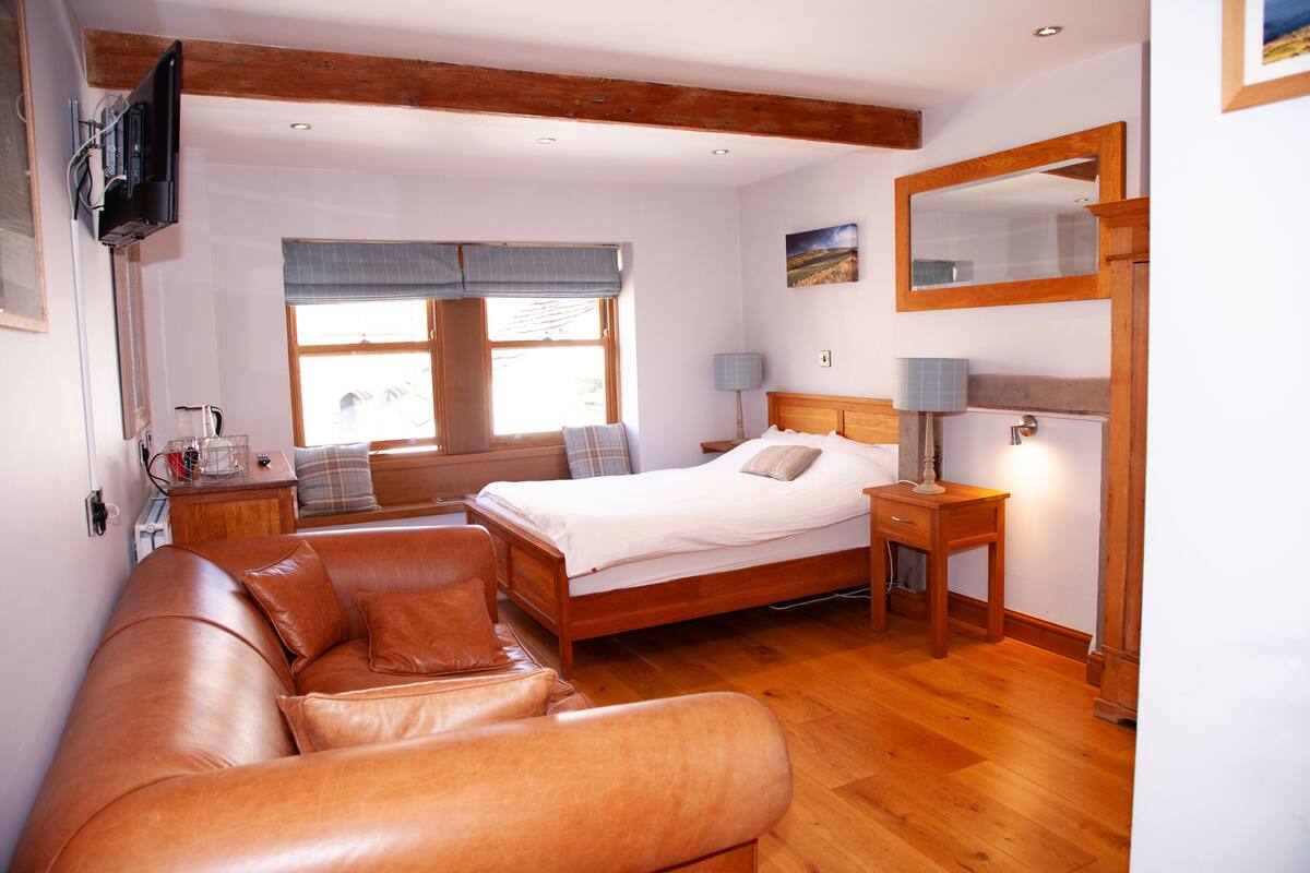 A bedroom with a bed, a wooden headboard, a bedside table with a lamp, a wooden bench, and a television mounted on the wall.