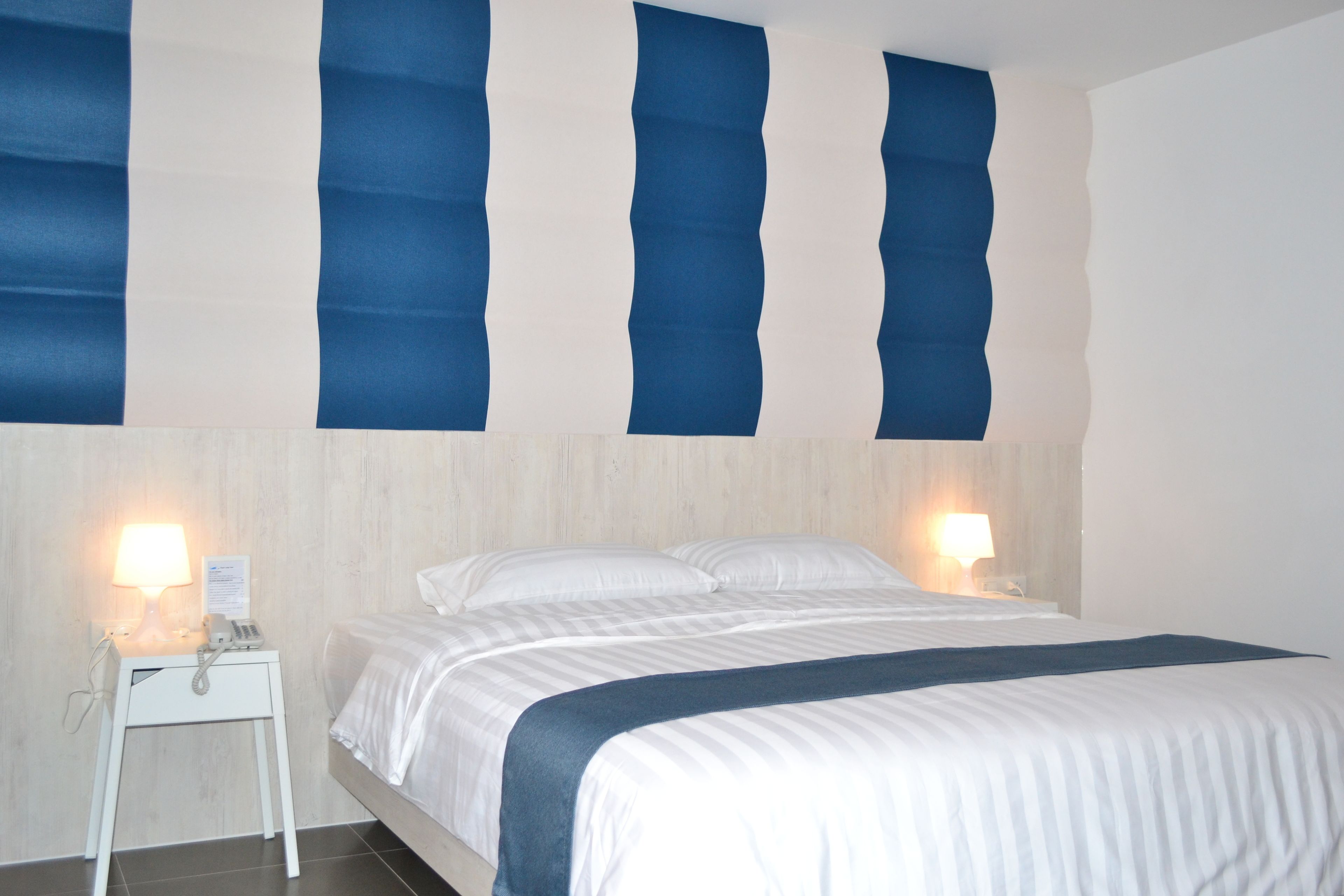 A hotel room with a bed, two bedside lamps, a nightstand, and a wall with vertical blue panels.