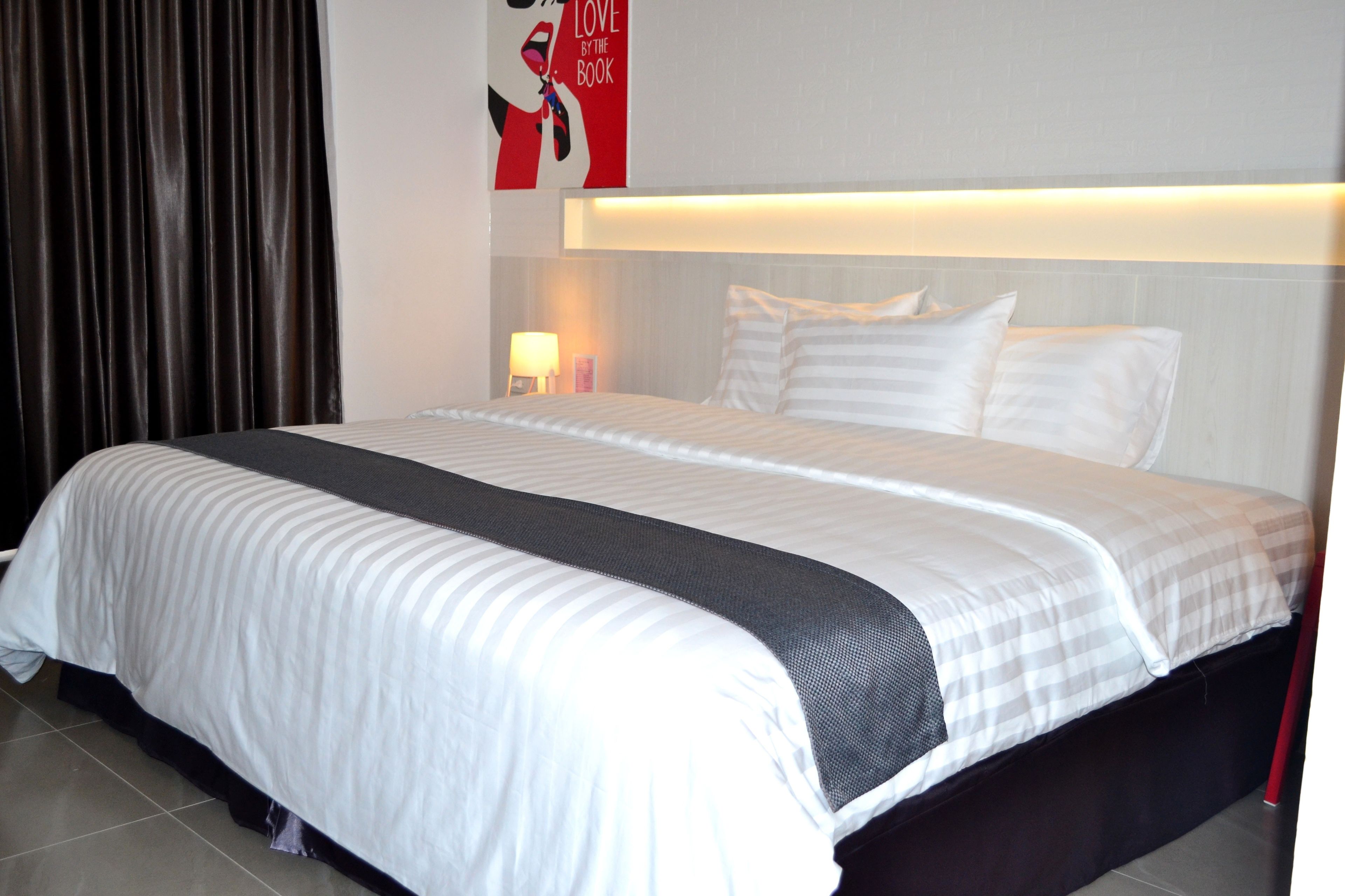A neatly made bed with white and gray bedding, a dark headboard, and a wall-mounted light fixture.