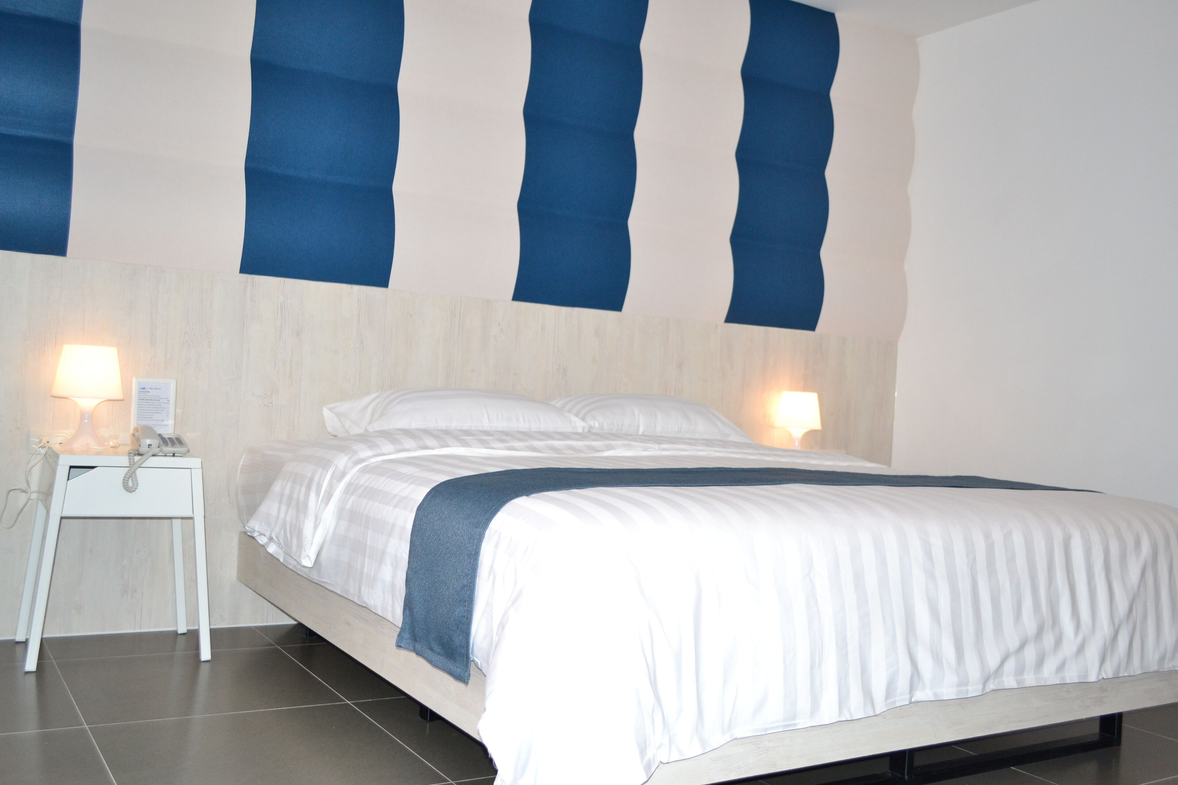 A neatly made bed with a white and blue striped blanket, a white bedside table with a lamp, and a wall with blue vertical stripes.