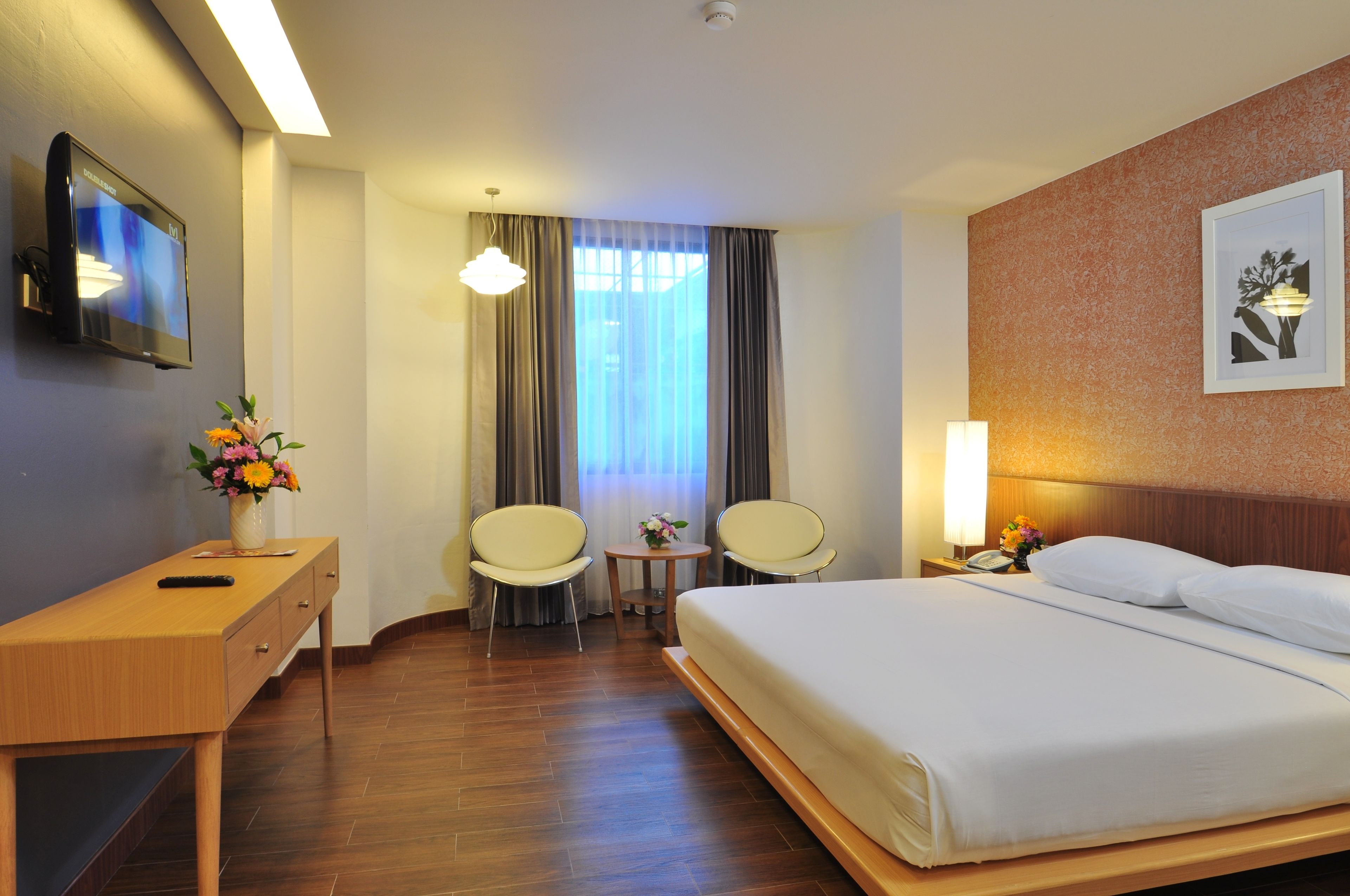 A hotel room with a bed, a desk, two chairs, a small table with a vase of flowers, a TV, and a framed picture on the wall.
