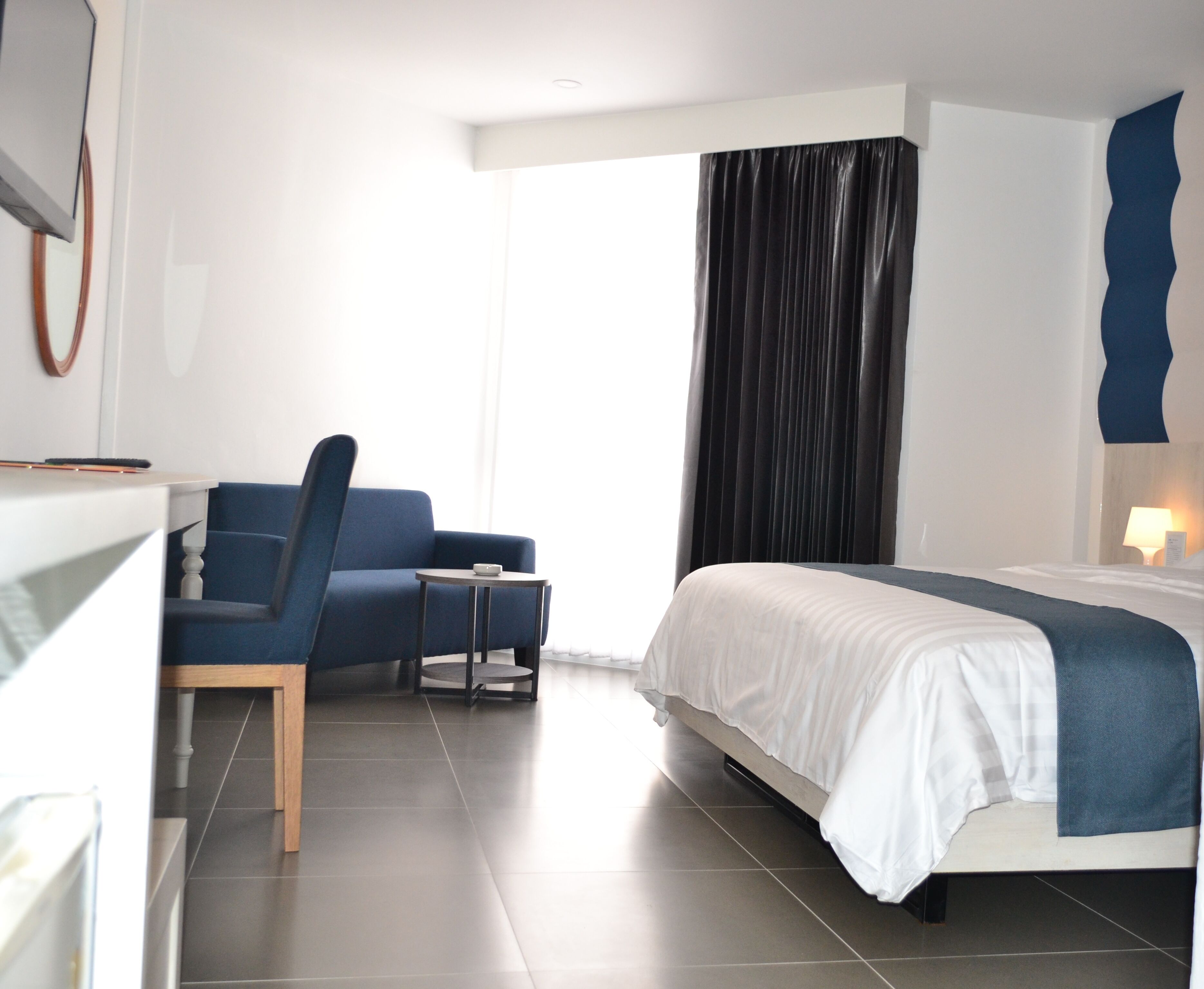 Una camera d'albergo moderna con un letto, una poltrona blu, un tavolino e un'ampia finestra con tende.