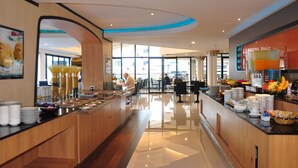 Um lobby de hotel moderno com um balcão de recepção comprido, uma área de buffet com diversos alimentos e uma área de estar com mesas e cadeiras.