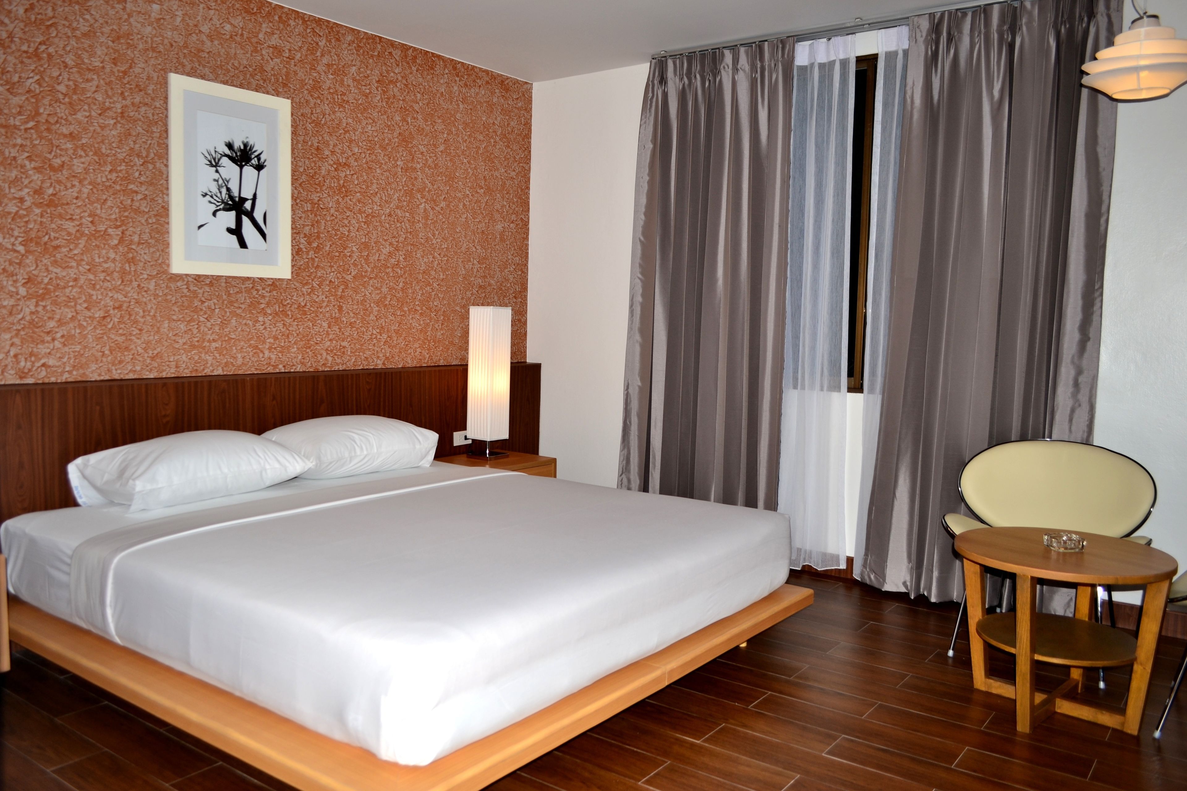 A hotel room with a large bed, a bedside table with a lamp, a chair, and a window with curtains.