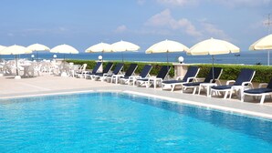 Área à beira da piscina com espreguiçadeiras e guarda-s óis, céu claro e vista para o oceano.