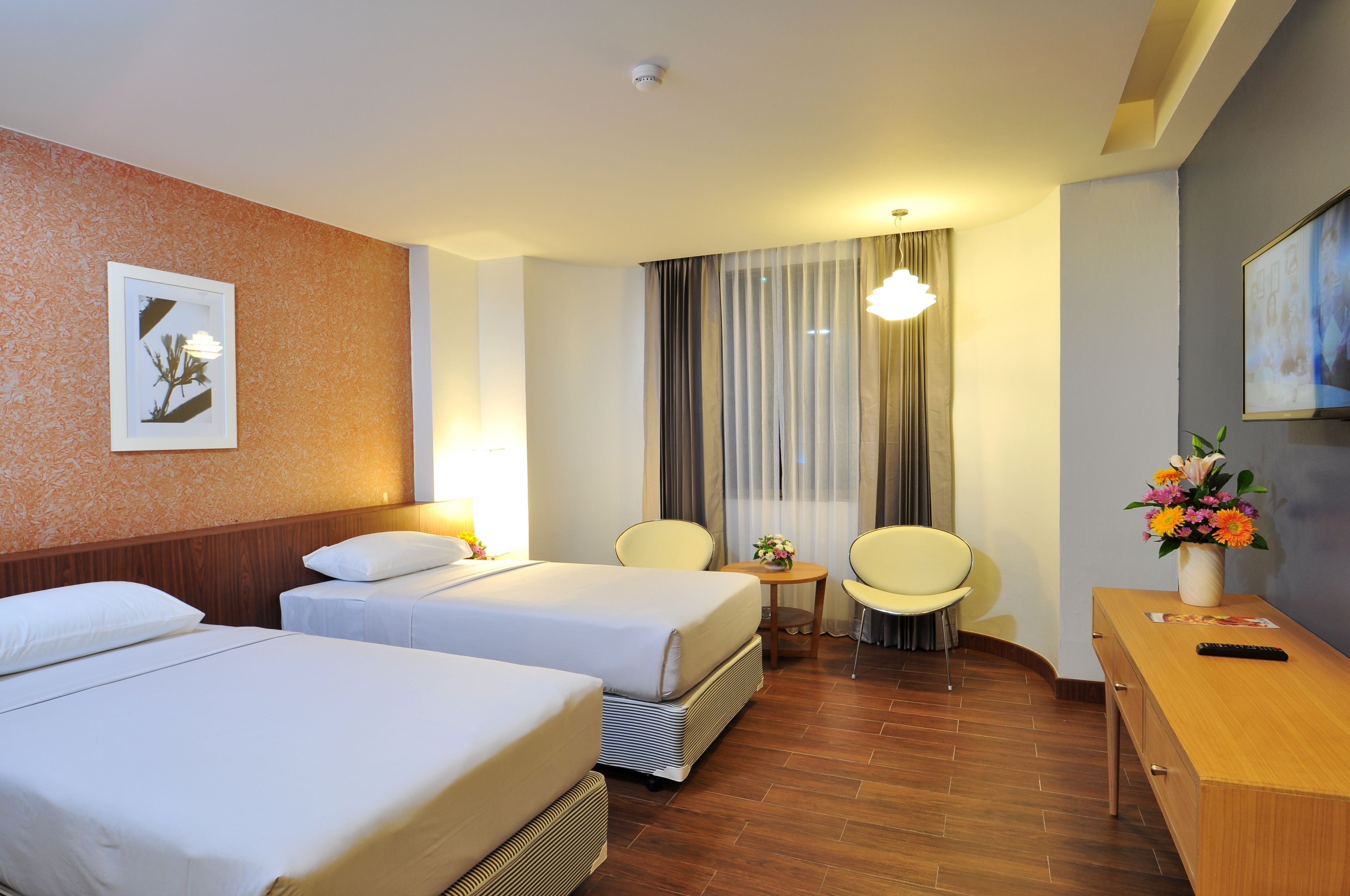 A hotel room with two beds, a small table with a vase of flowers, a chair, and a desk with a remote control.