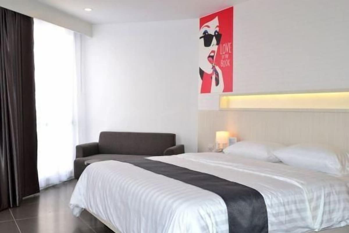 A hotel room with a bed, a chair, a poster on the wall, and a window with curtains.