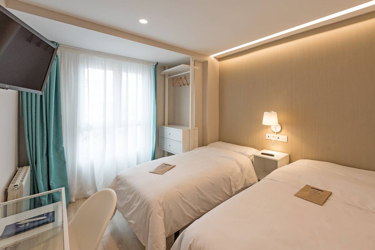 A hotel room with two beds, a desk, a TV, and a window with curtains.