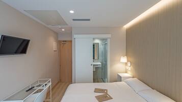 A modern hotel room with a bed, a desk with a chair, a TV mounted on the wall, a bedside lamp, and a bathroom visible through a glass door.