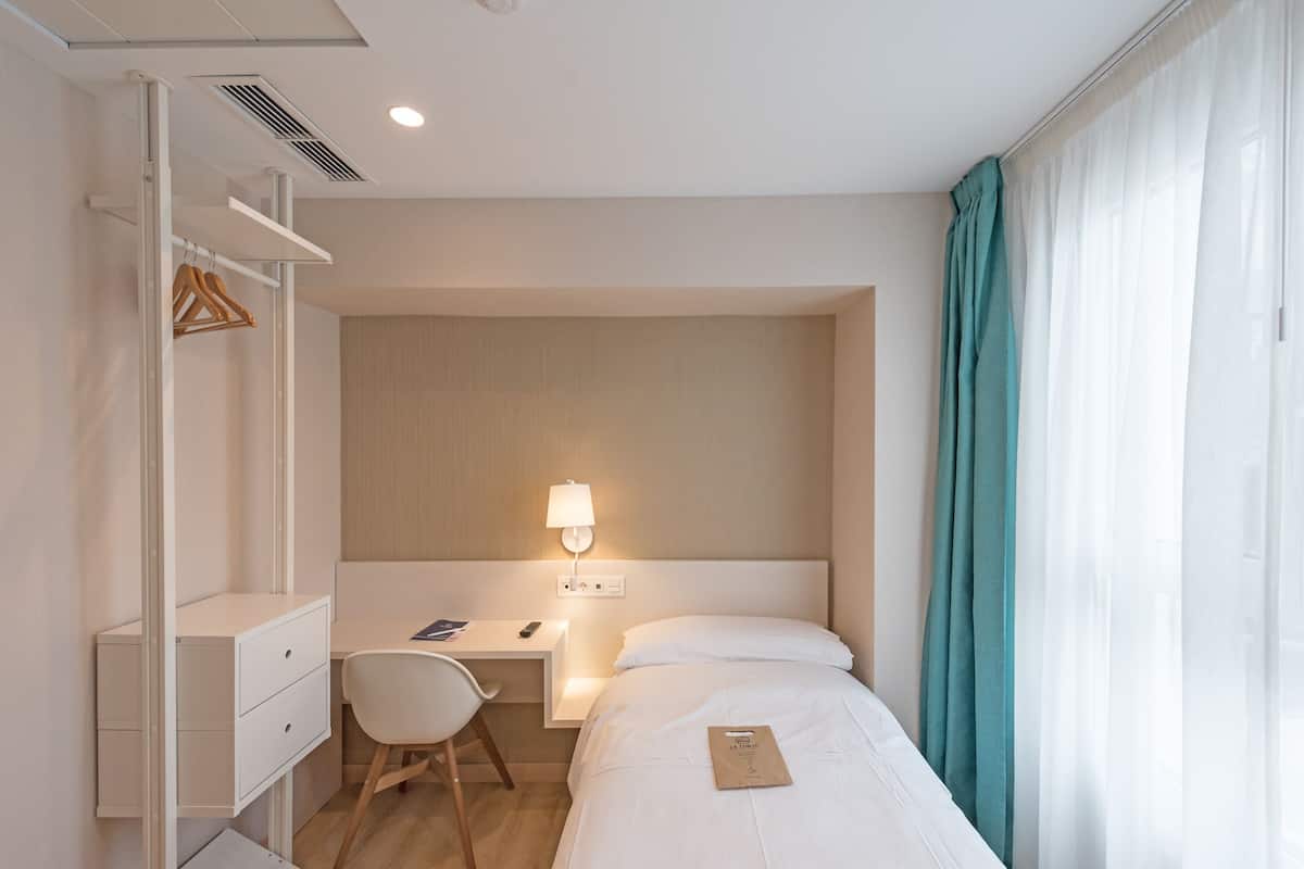 A hotel room with a bed, a desk with a chair, a wardrobe, and a bedside lamp.