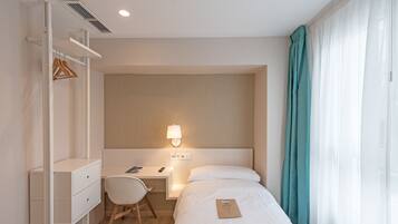 A hotel room with a bed, a desk with a chair, a wardrobe, and a bedside lamp.
