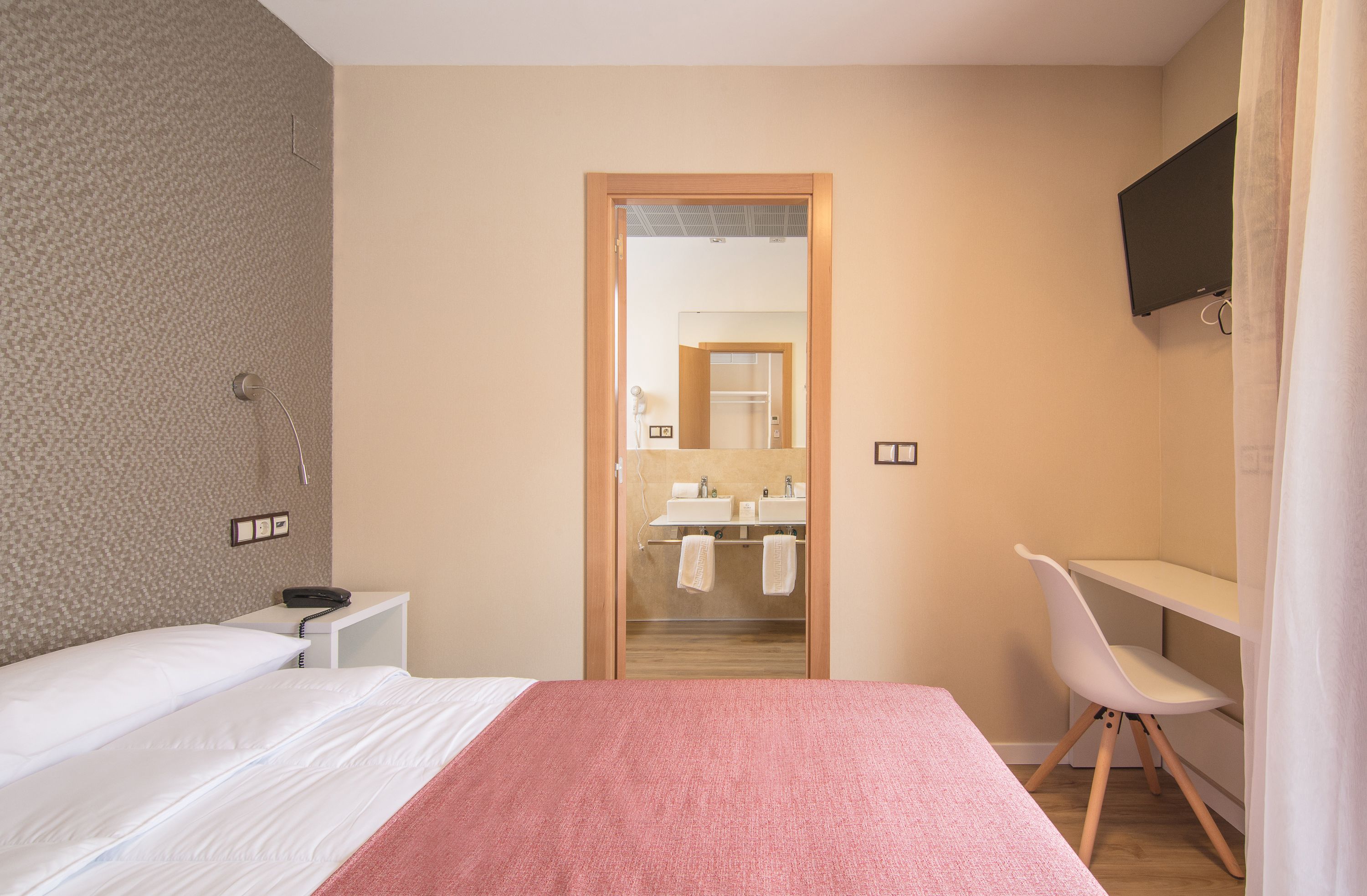 Habitación de hotel con cama, escritorio, silla, televisor y baño visible a través de una puerta abierta.