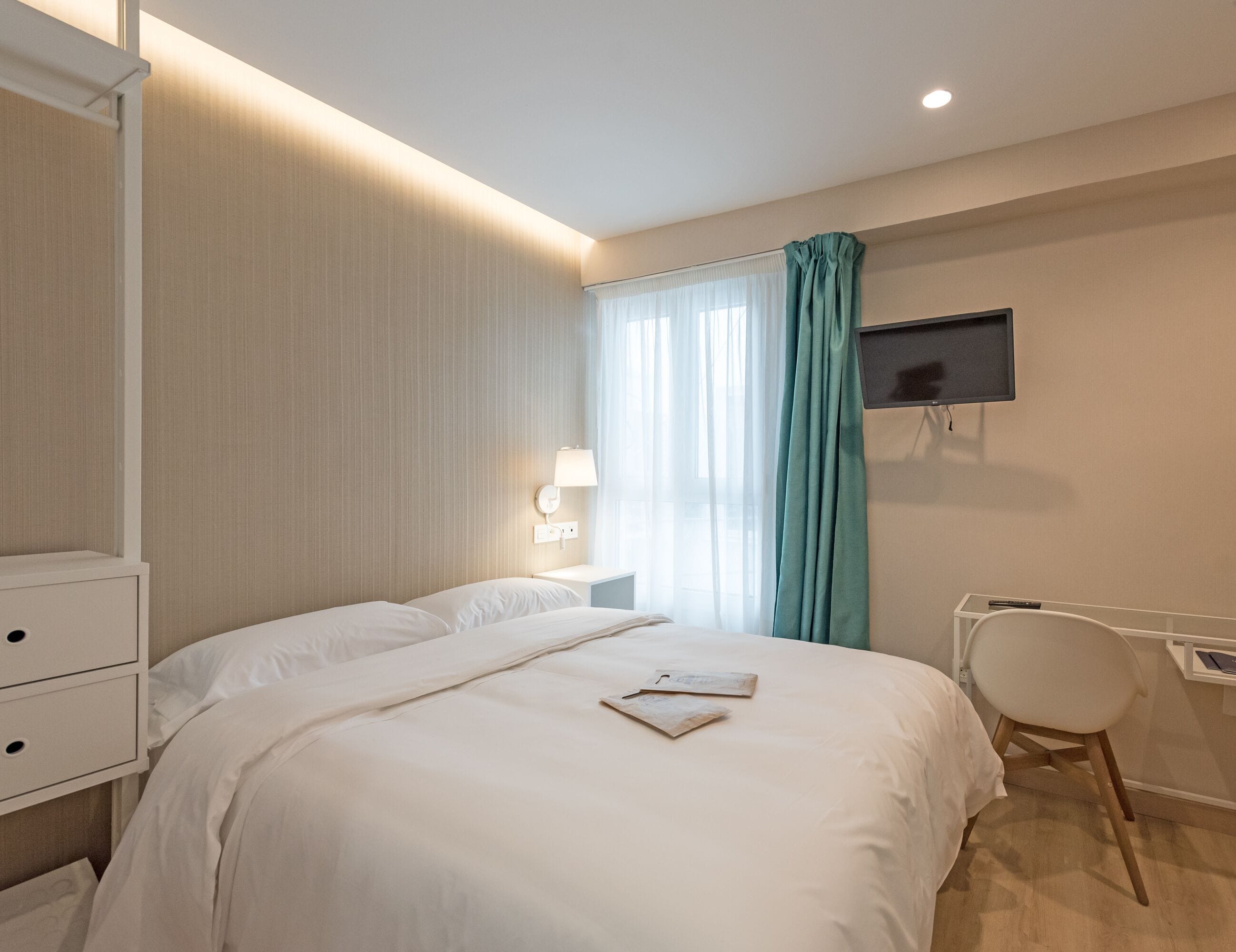 Een hotelkamer met een bed, een bureau met stoel, een aan de muur bevestigde televisie en een raam met gordijnen.