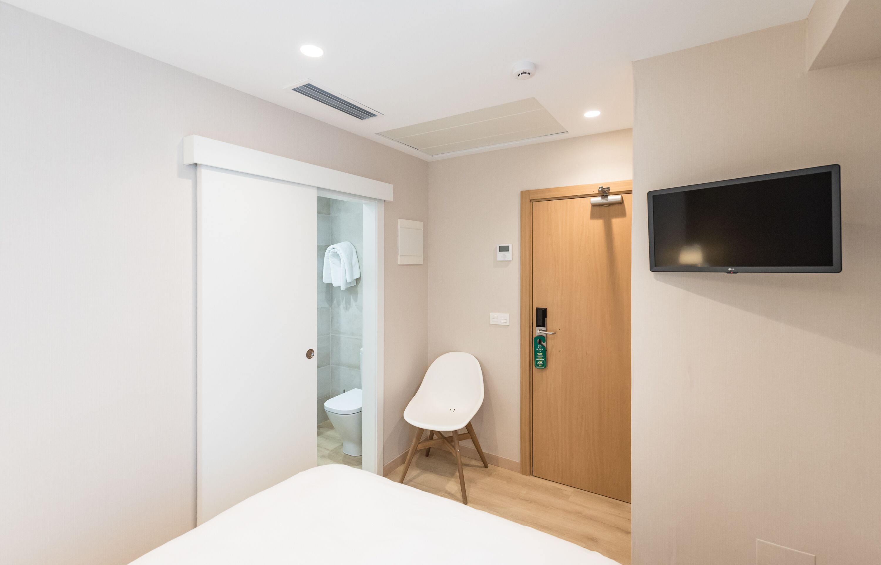 A hotel room with a bed, a chair, a television, a wooden door, and a bathroom visible through a glass door.
